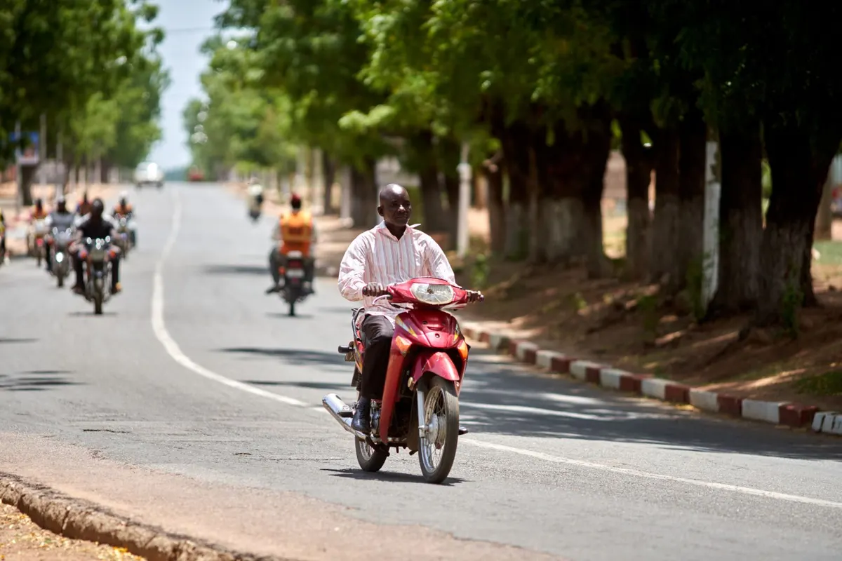 Man riding a red motorbike down a sunny tree-lined street